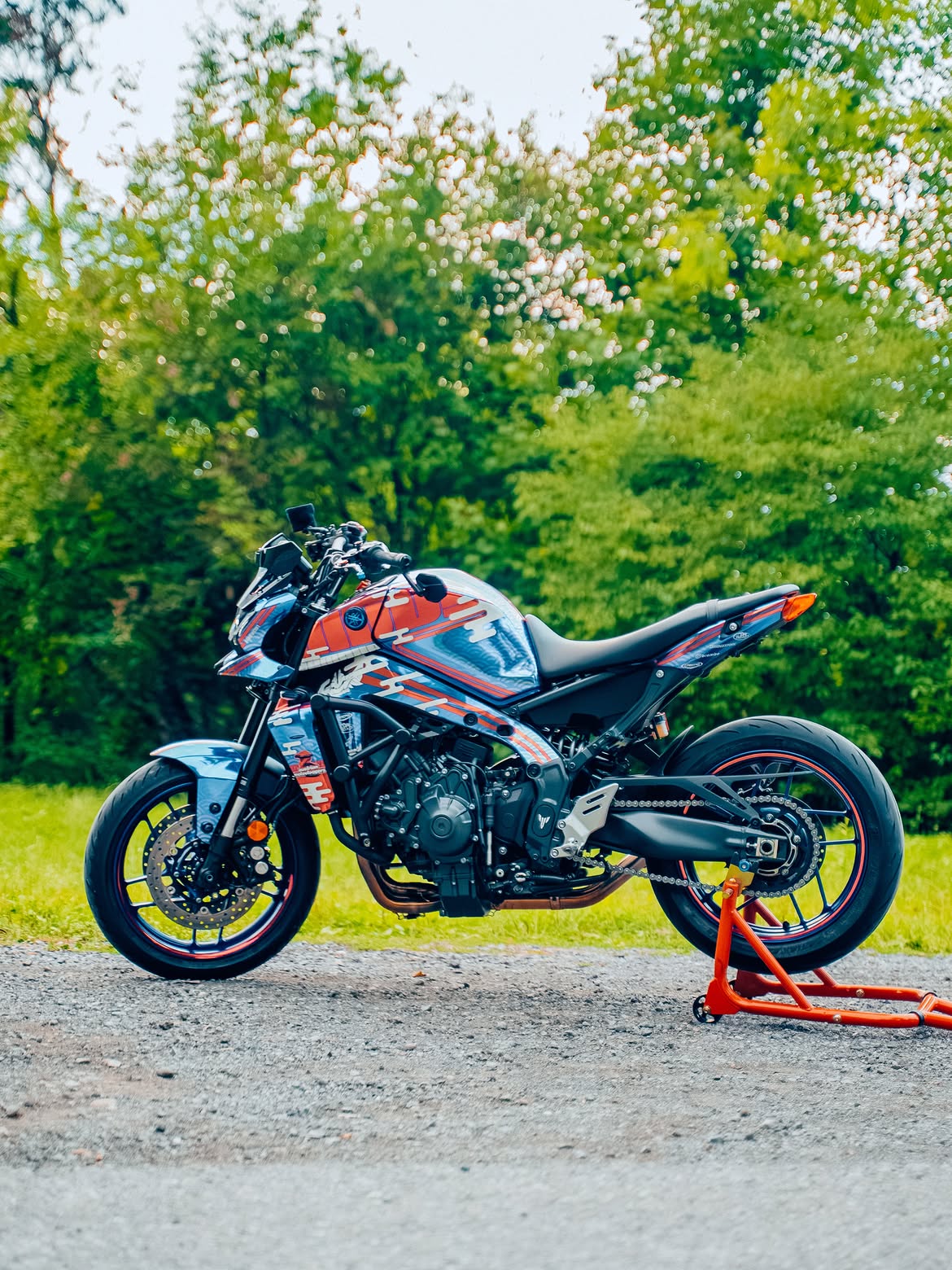 A custom blue, red, and white motorcycle with racing decals is parked on a gravel surface on a red stand, with a lush green forest background.