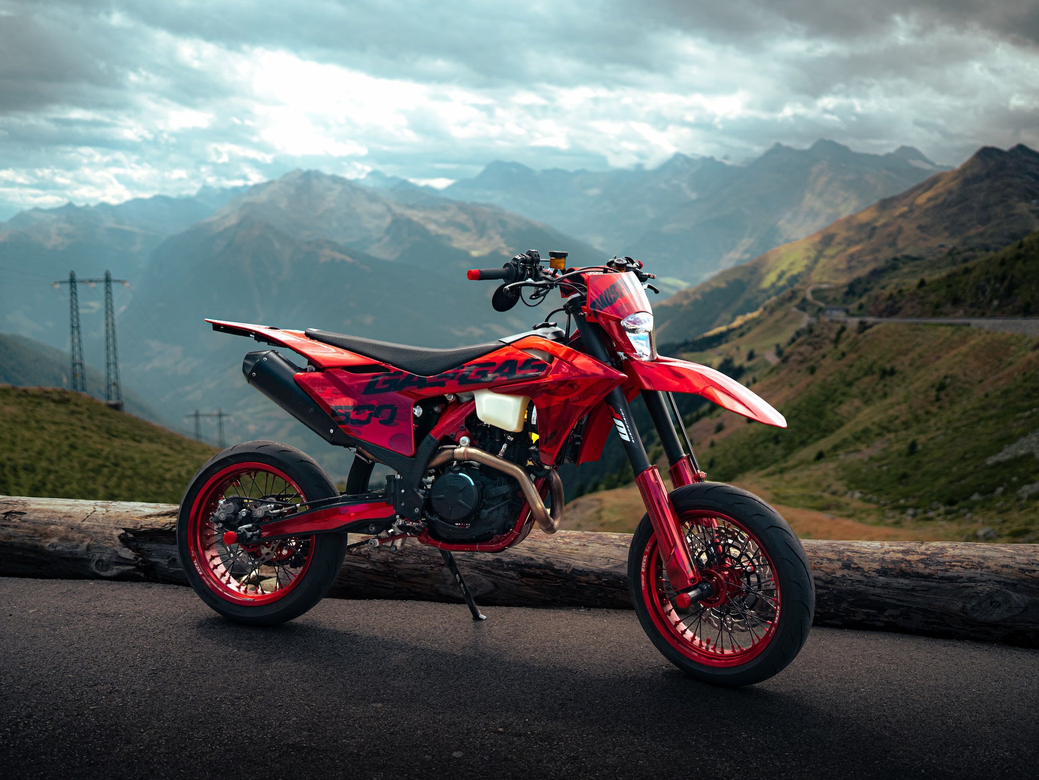 A red and black dirt bike motorcycle is parked on an asphalt road with a large log in the foreground, set against a dramatic mountain landscape under a cloudy sky.